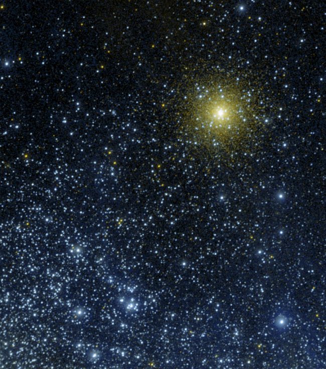 Small, hazy yellowish star cluster on the outskirts of a larger group of blue stars.