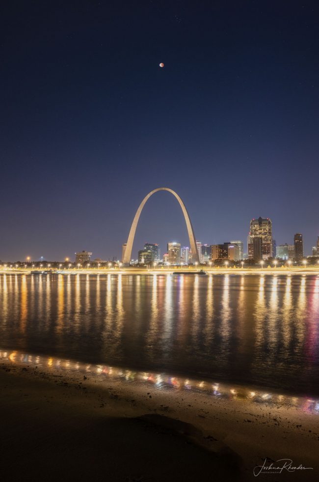 Giant St. Louis arch with city lights reflected in river and eclipsed moon above.
