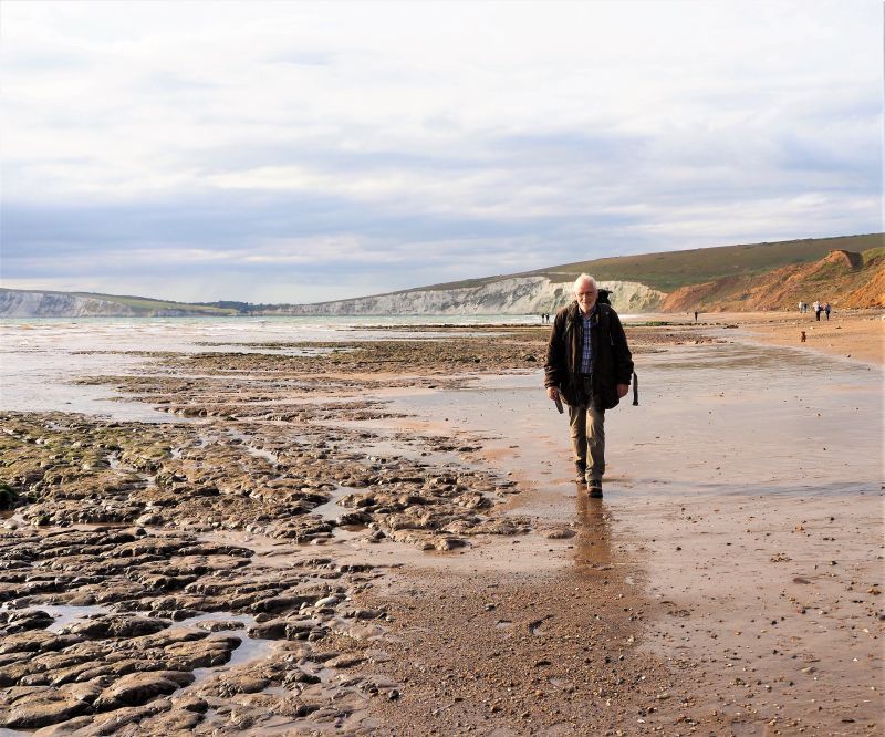 Man walking on wet sand next to large inlet, with cloudy sky and hills in background.