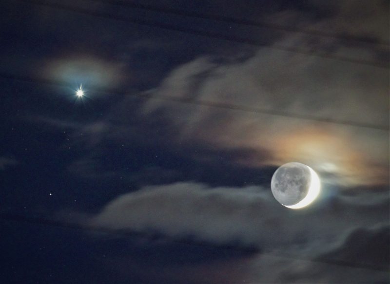 Crescent moon with earthshine near Venus, shining between fuzzy clouds.