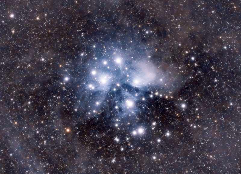 Bright stars at center and blue-gray cloudiness surrounding in extremely dense star field.
