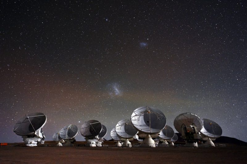 11 dish telescopes in front of starry background with 2 small, fuzzy, glowing patches in the sky.