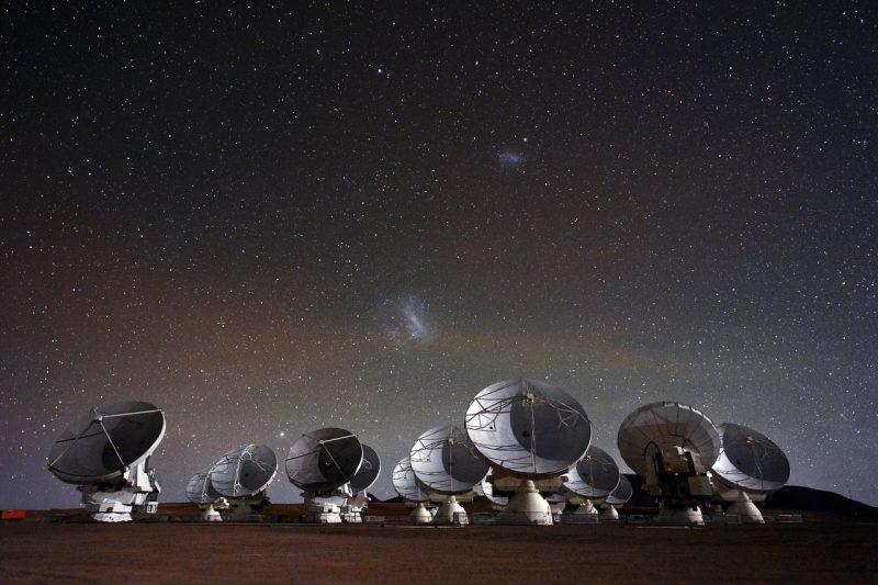 Dish telescopes in front of starry background with one large cloudy smear and one smaller smear above.