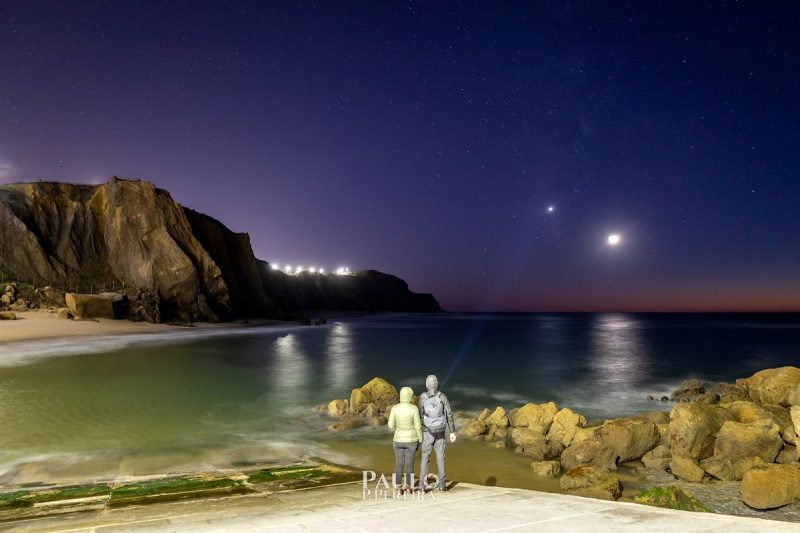 Two people standing in front of sea, looking out at moon and Venus.