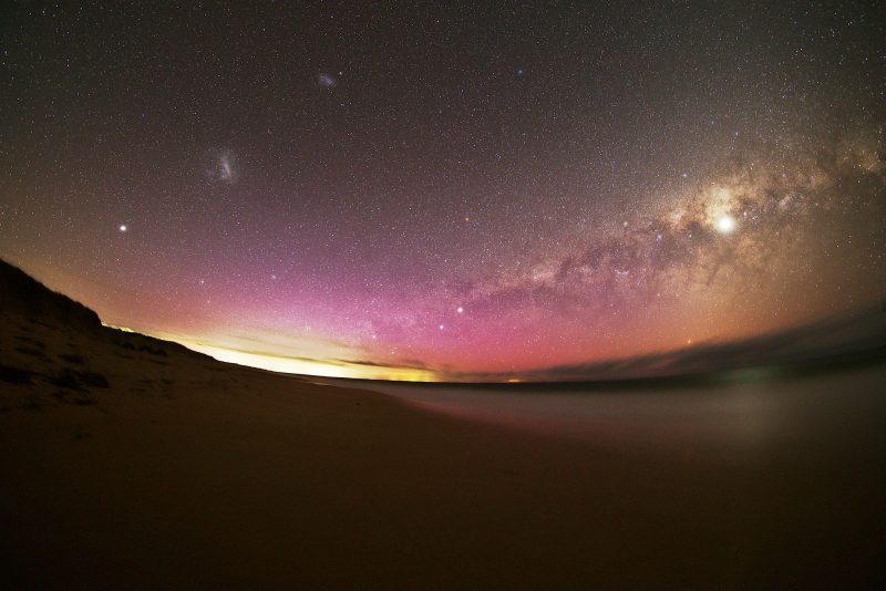Red and yellow glow along horizon with cloudy Milky Way and Small and Large Magellanic Clouds.