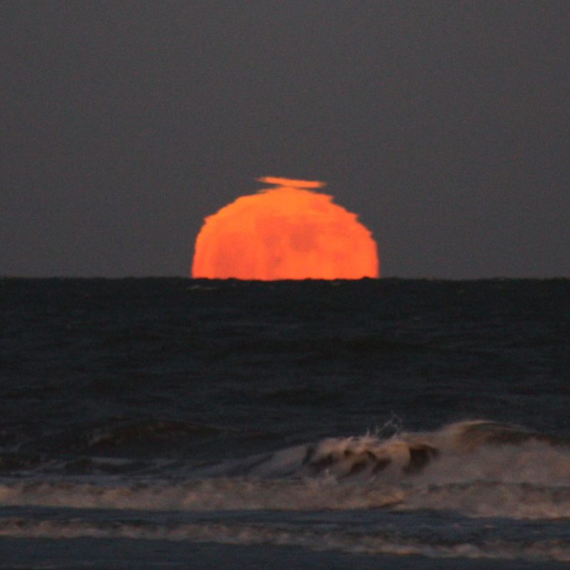 Oddly shaped suns and moons: Top half of a wavering orange moon rising over ocean.