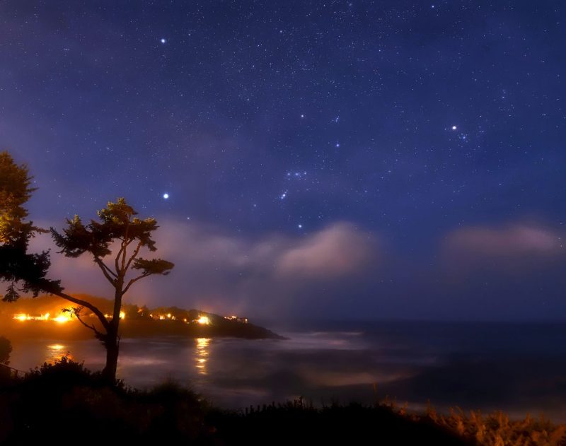 Misty view of Orion and Sirius in deep blue sky, with clouds below and town lights on a shore.