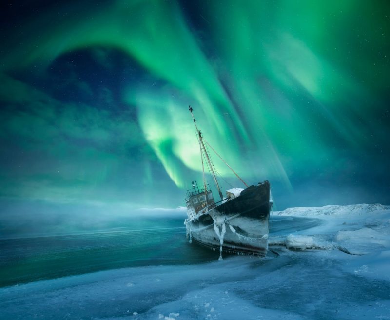 Green swirls of northern lights over a dark blue sky with an icy ship stuck on shore.