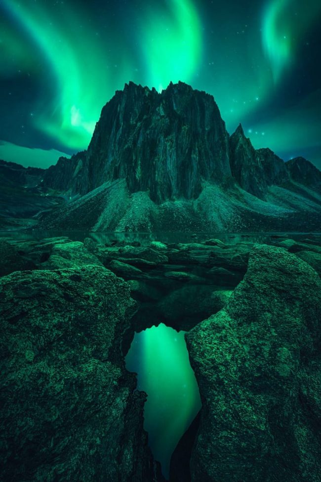 Large rocky mountain in front of green tentacles of light reflected in foreground pool.