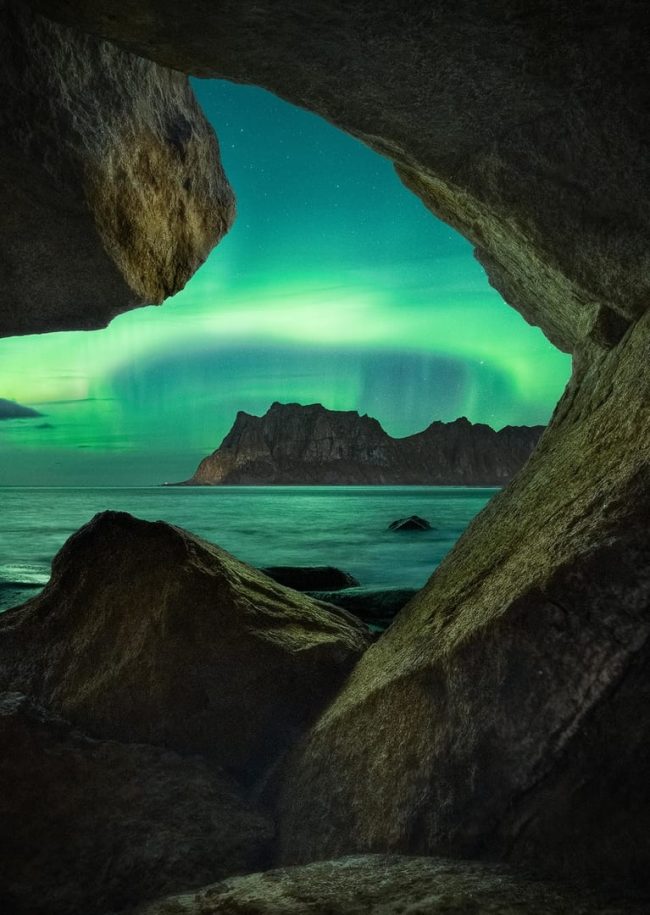 A ring of green aurora over cliffs and water seen through a rocky cave.