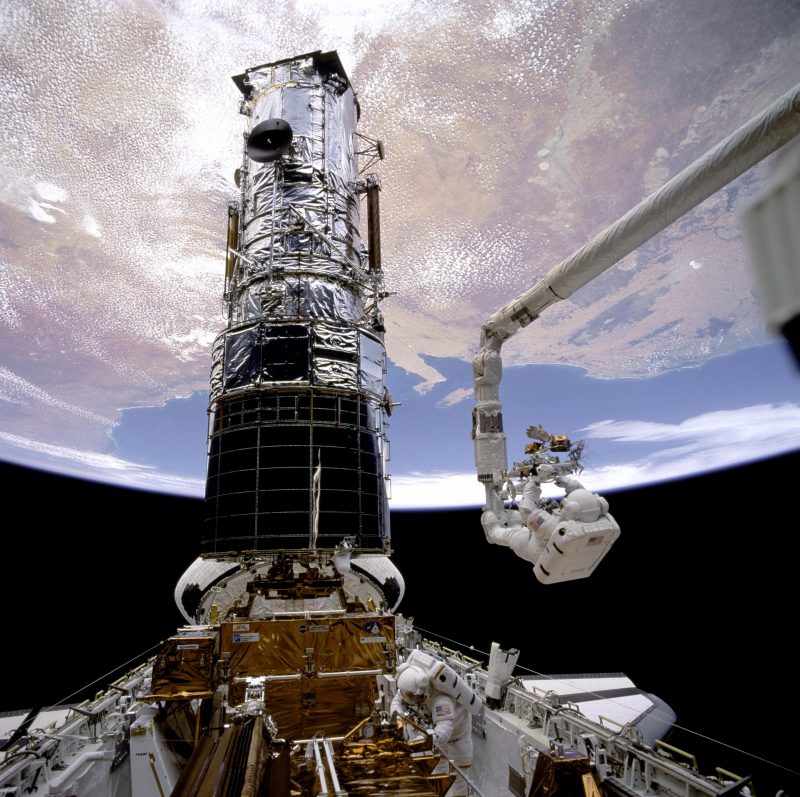 Huge metallic structure floating in space with an astronaut repairing a piece. Earth is in the background.