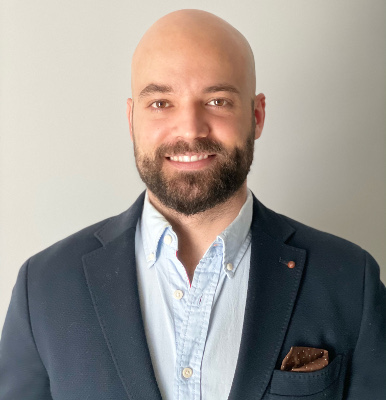 Bald man with dark beard and mustache in front of white wall.