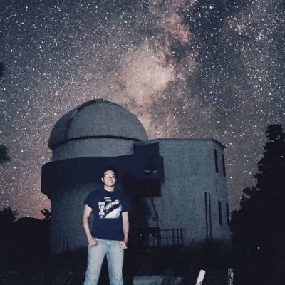 Man in t-shirt stands in front of observatory with Milky Way behind.