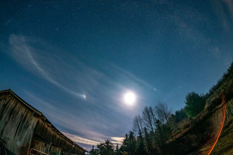 Fisheye view of bright moon and streaky clouds with a rainbow patch and some stars.