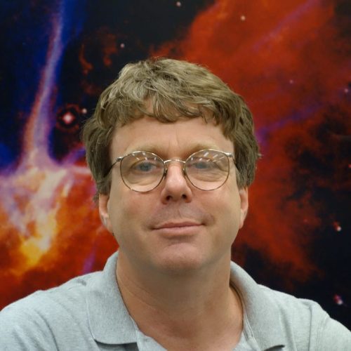 Smiling man with eyeglasses and space nebula mural behind him.