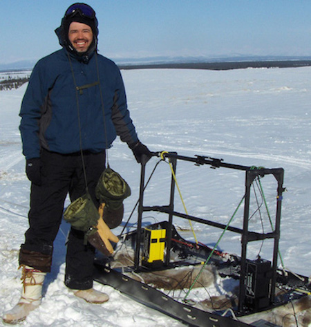 Smiling man standing next to sled on snow-covered ground.