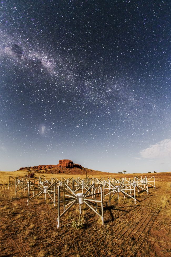 Milky Way over red rocky landscape with metal spider-like structures in foreground.