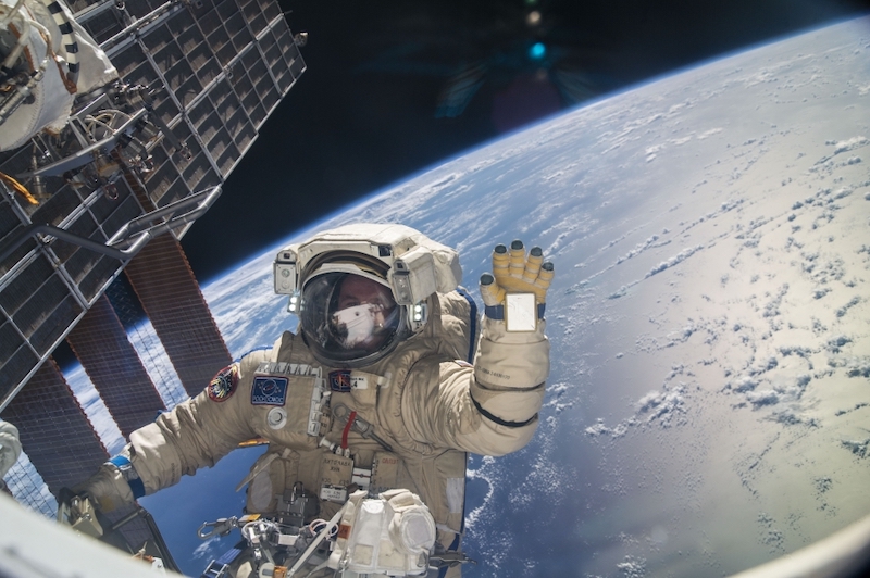 2022's 1st spacewalk: A man wearing a white astronaut suit with red stripes waves to the camera. A blue Earth and the blackness of space behind him set the backdrop.