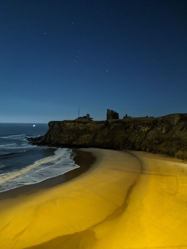 Night photo: yellow beach with ruins on promontory and constellation Orion above the sea.