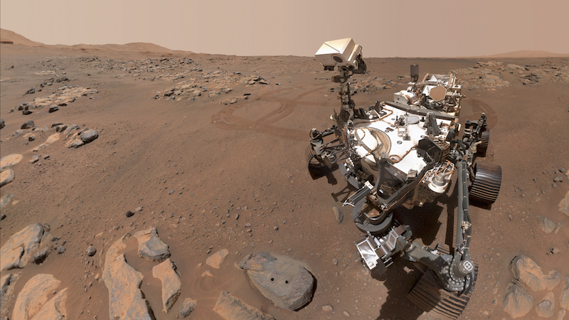 Mechanical rover on reddish terrain with reddish sky.