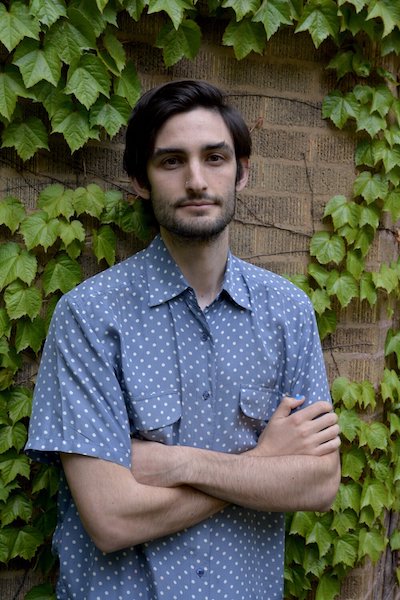Serious, bearded young man in polka-dotted shirt with his arms crossed in front of ivied wall.