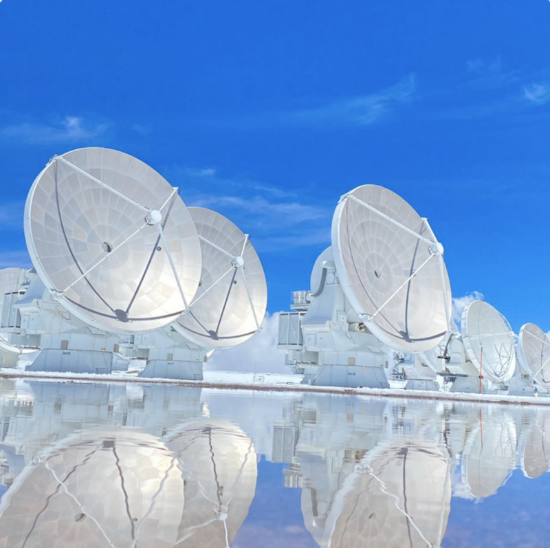 Radio dishes under a blue sky, standing in a glittering pool of water.