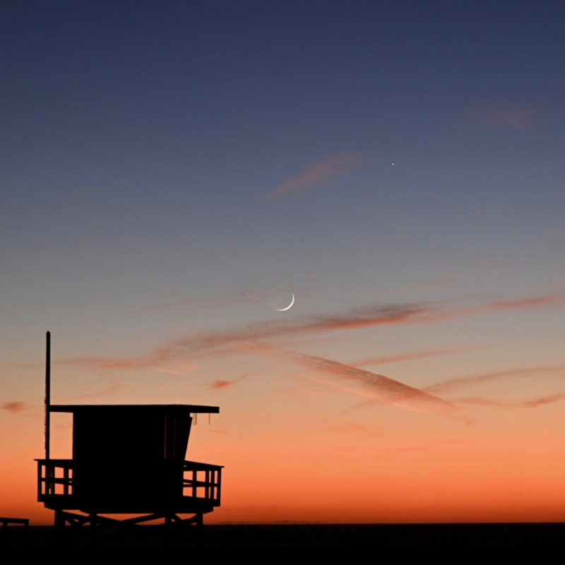 Silhouette of lifeguard tower in the foreground, crescent moon and Mercury in a twilight sky.
