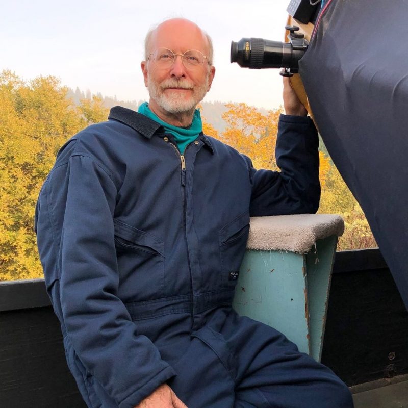 Don Machholz: Bald man with white beard standing next to a large telescope.