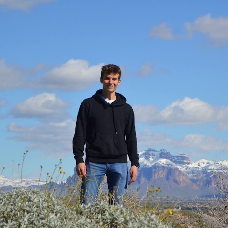 Young man standing in field with mountains and puffy clouds behind him.