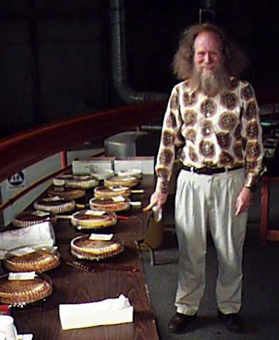 Man with long hair standing next to table with pies on it.