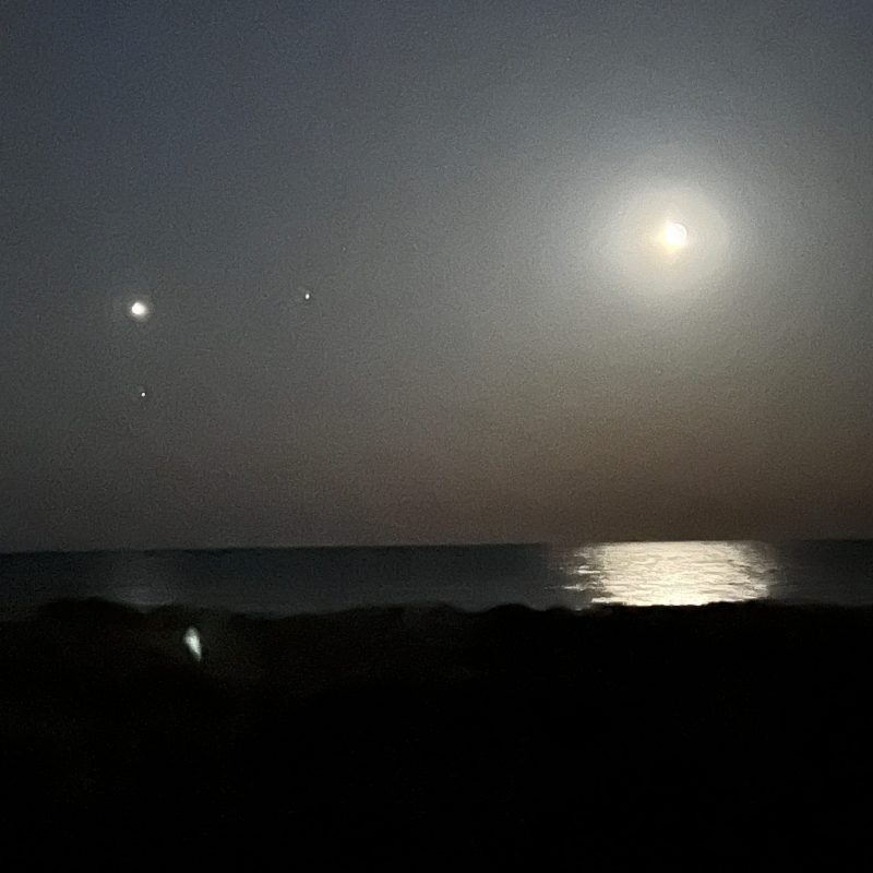 A beach at night with the moon on the right side and its reflection in the water. Planets on the left.