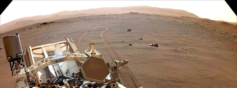 Perseverance rover: brownish terrain with part of rover in foreground and hills in background.