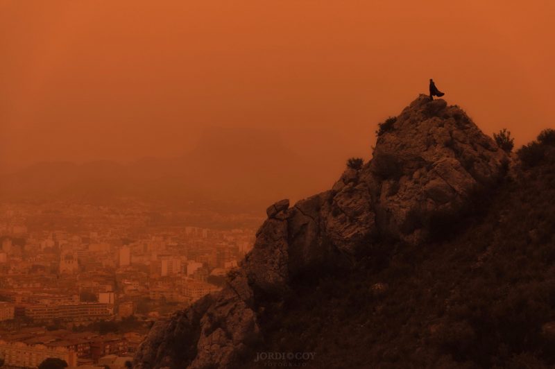 Orange dusty sky with mountain in front and person standing atop.