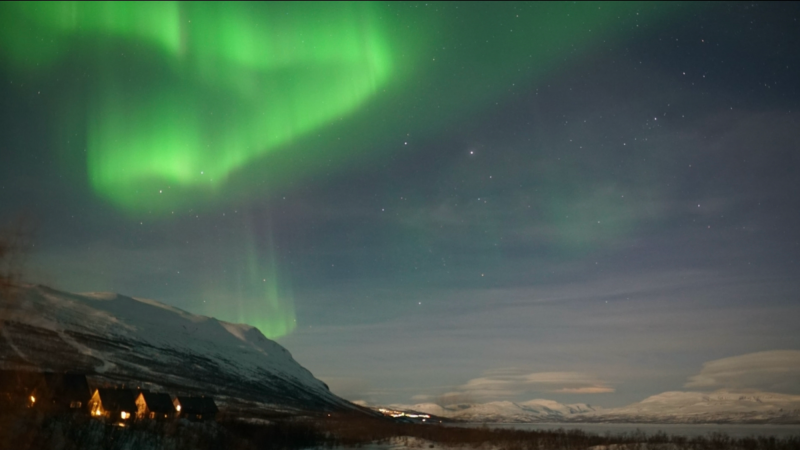 Aurora season: Green aurora above hilltops, with a body of water to one side.