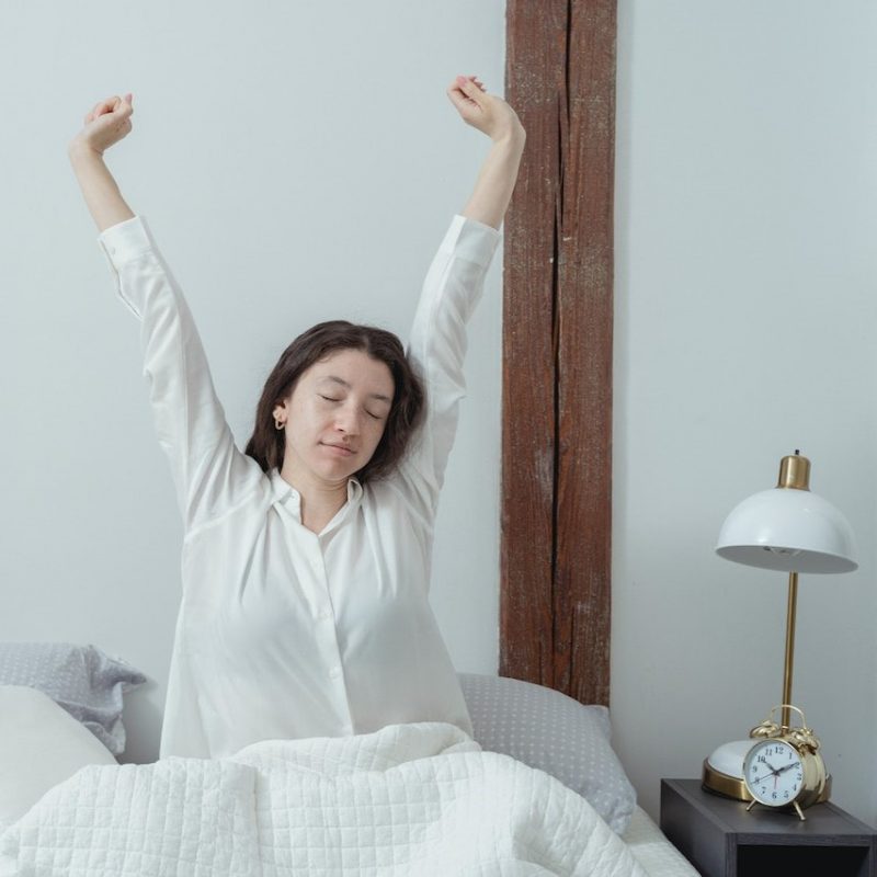 Woman wakes up and stretches with alarm clock by bed.