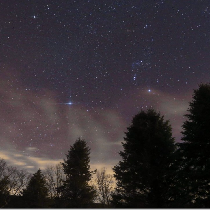 Starry, dark sky with the very bright, bluish stars from the constellation Orion and a bright star, Sirius, with 4 streaks coming out of it.