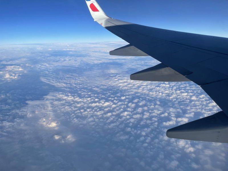 The wing of the aircraft above a layer of small clouds and a blue sky over the distant horizon.