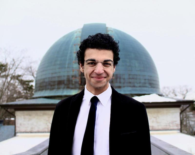 Smiling man with dark curly hair in suit and tie standing in front of a large domed building.