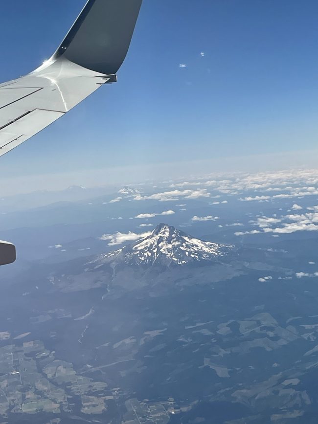 Plane wing at top corner and snow-capped mountain with some puffy clouds and darker haze.