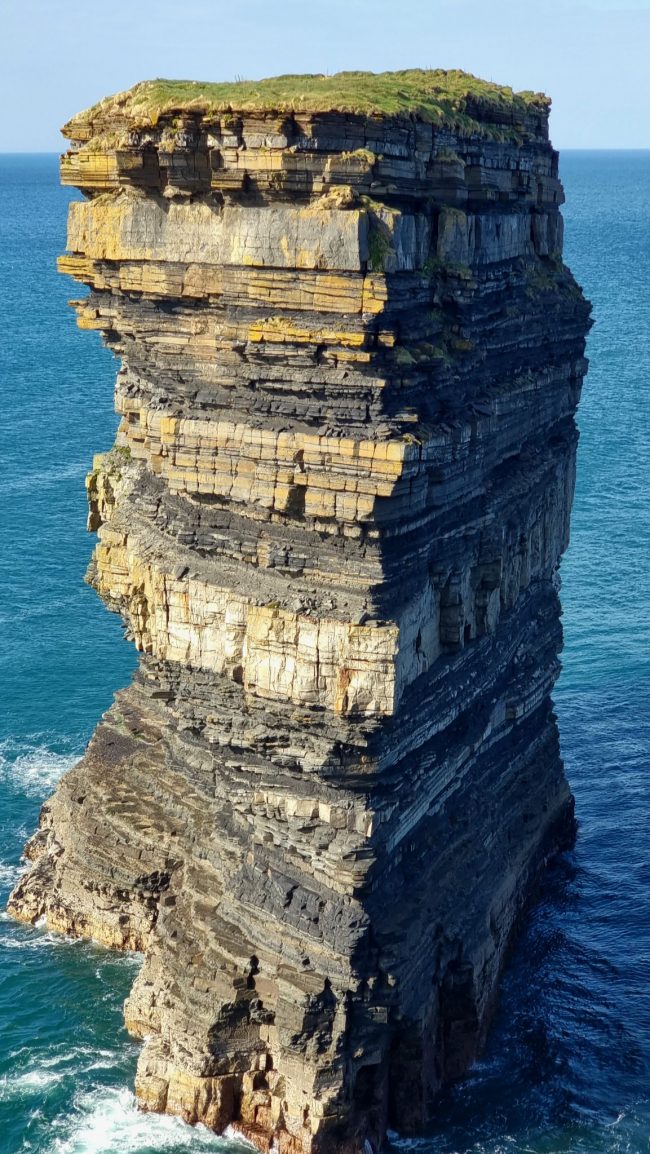 Sea stack with many layers surrounded by blue water and waves.