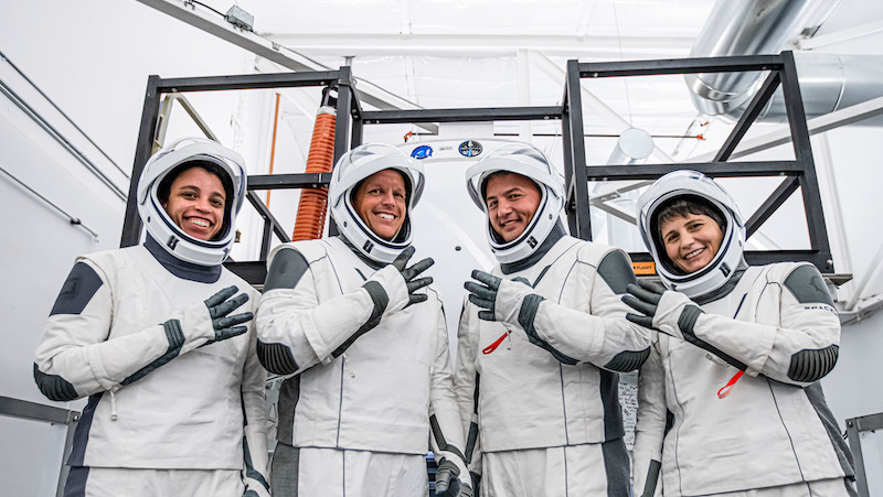 Crew-4; Four people dressed in white spacesuits are standing in a white room with metal bars behind them. They’re all smiling and holding up four fingers each.