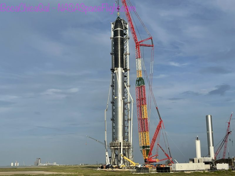 Starship: Cylindrical rocket with silver lower part and black upper part, next to red and yellow crane.