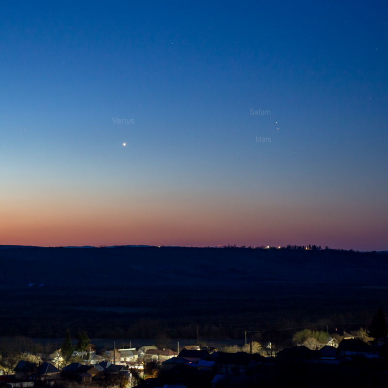 Venus, Saturn and Mars are close in a orange and blue sky during sunrise over lighted village.