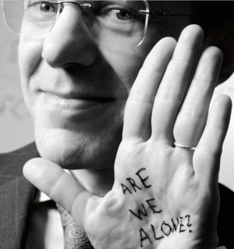 Closeup of man in glasses with hand by face and 'Are we alone' written on palm with marker.