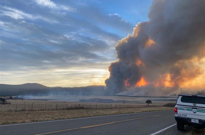Giant clouds of smoke rising into blue sky from barely huge fire viewed from a highway.
