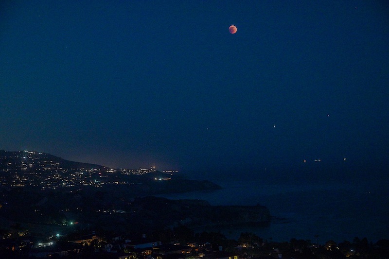 Tiny red moon in deep blue sky over lights of towns along a coastline.