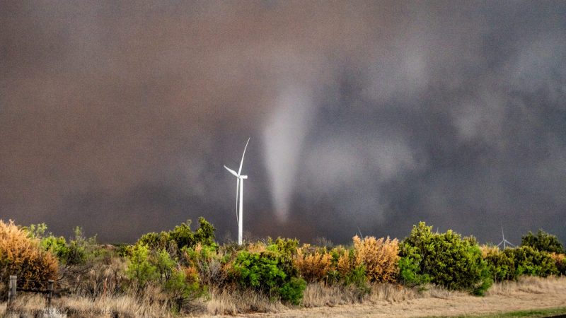 Brown and white clouds with funnel kicking up dust, tall white windmill in front.