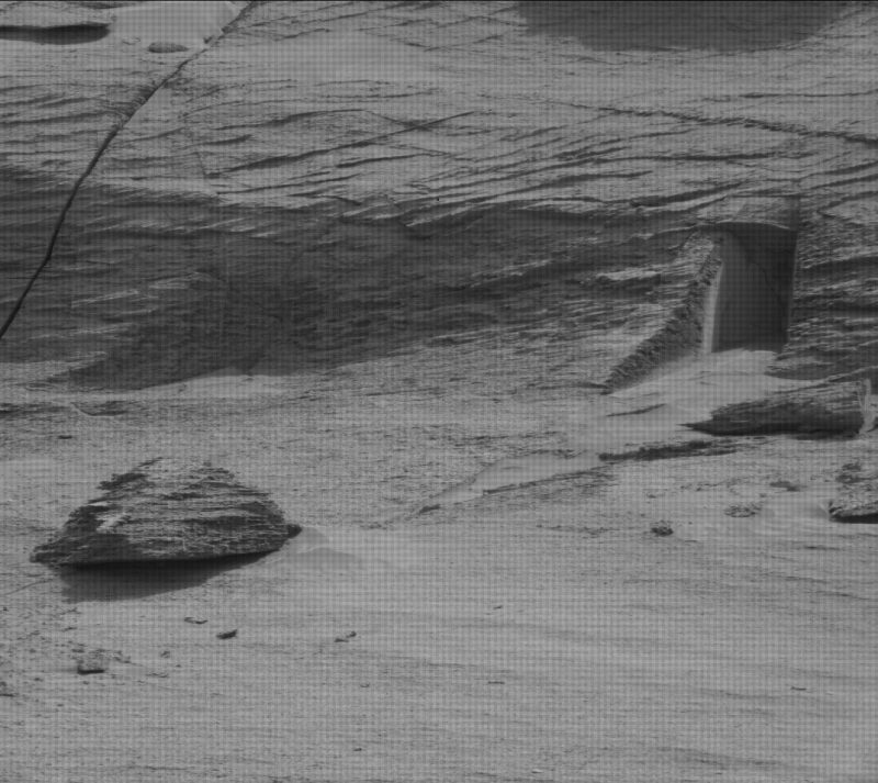 Sandy landscape with rock at left and rectangular-looking doorway at right in steep rocky bank.
