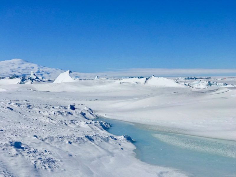 Below Antarctic ice: Clear blue sky and a mountain with snow and a clear water river.