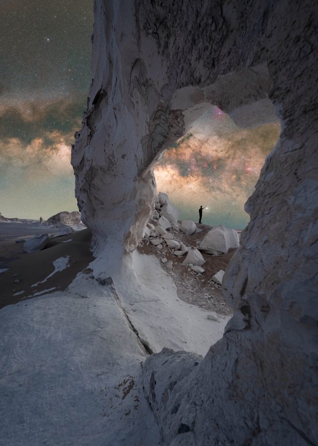Milky Way appears through an irregular rock arch with a man in the distance.